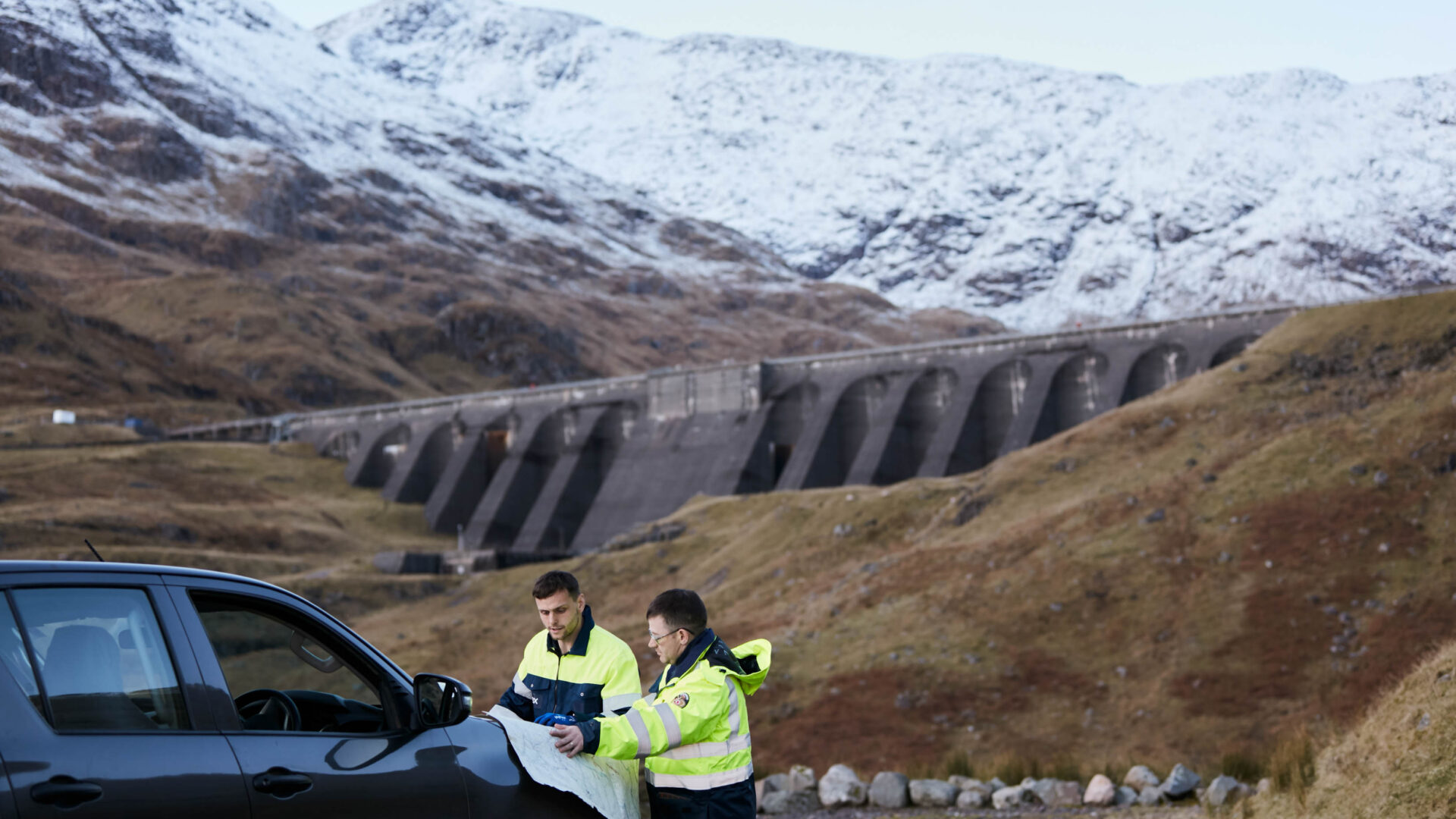 Two Drax workers looking at documents by a car, in front of Cruachan Power Station dam
