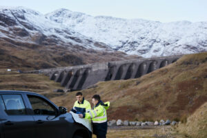 Two Drax workers looking at documents by a car, in front of Cruachan Power Station dam
